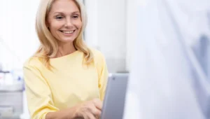 A smiling woman with blonde hair wearing a yellow top is looking at and using a digital tablet, with a blurred person in a white coat visible in the foreground.