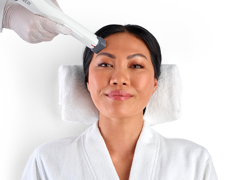 A woman in a white robe lies on a towel with her hair pulled back while a gloved hand uses a handheld device on her forehead, suggesting a facial or skin treatment—similar to the advanced care offered with Emsculpt Neo in Scottsdale.