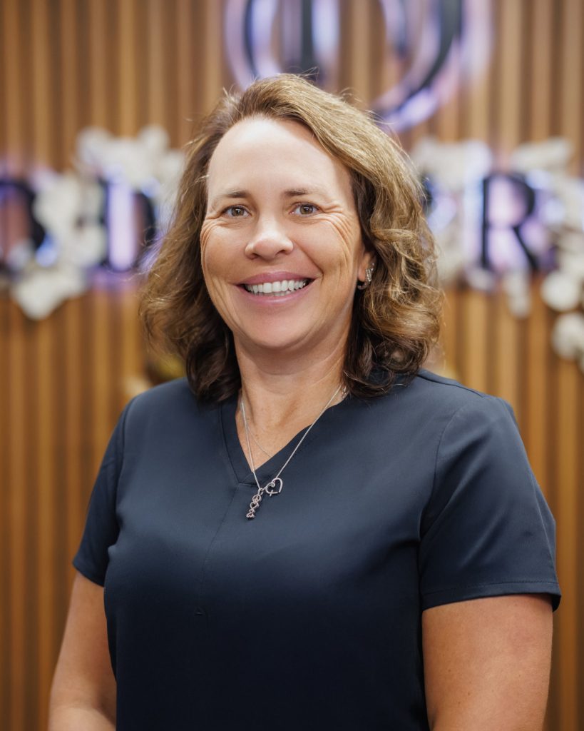 A woman with curly light brown hair wearing a navy blue top stands smiling in front of a wood-paneled background with blurred white signage and floral decorations, radiating confidence that could come from choosing the right dermal filler near me.